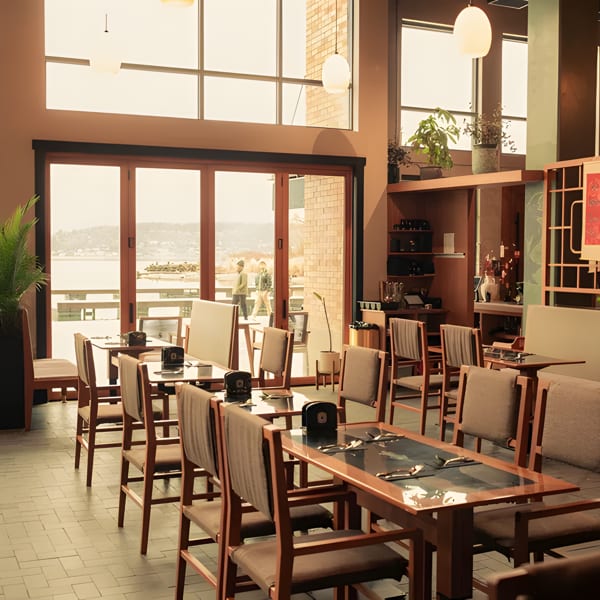 A restaurant dining room with wooden tables and chairs, large windows overlooking a body of water.