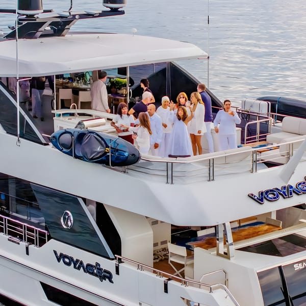 People socializing on the deck of a luxury yacht named "Voyager" on calm waters.