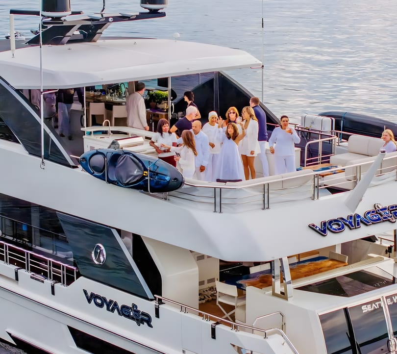 People socializing on the deck of a luxury yacht named "Voyager" on calm waters.