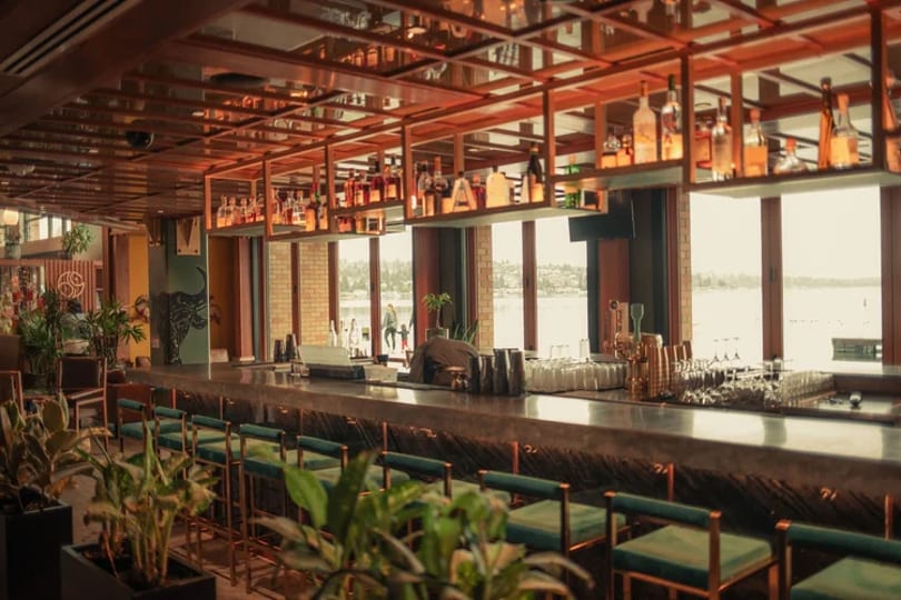 A modern bar with green stools, a long counter, and bottles on shelves, overlooking a lake.