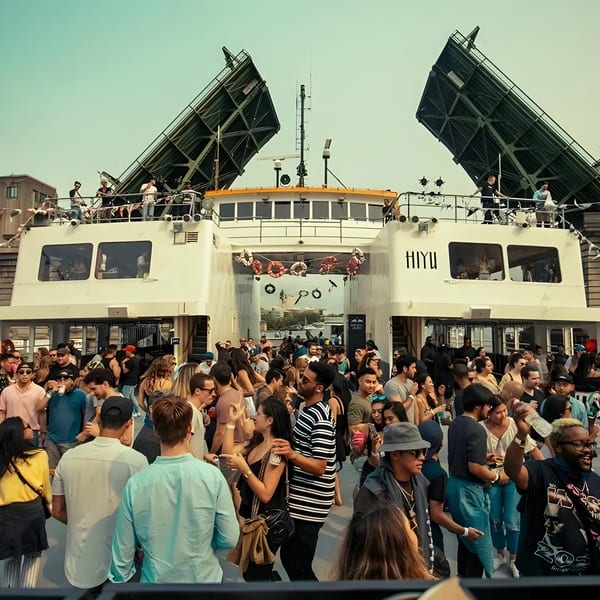 A lively crowd of people on a boat party, with a bridge in the background.