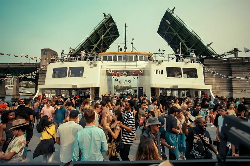 A lively crowd of people on a boat party, with a bridge in the background.