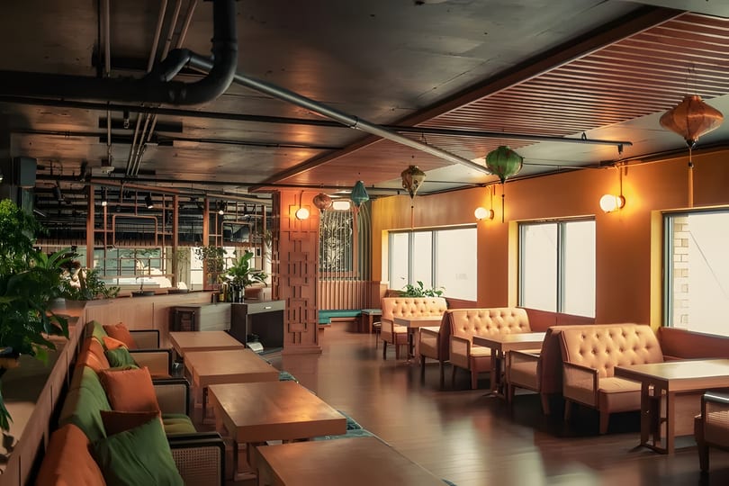A restaurant interior with orange walls, wooden tables, green and orange cushioned seating, and decorative lanterns.