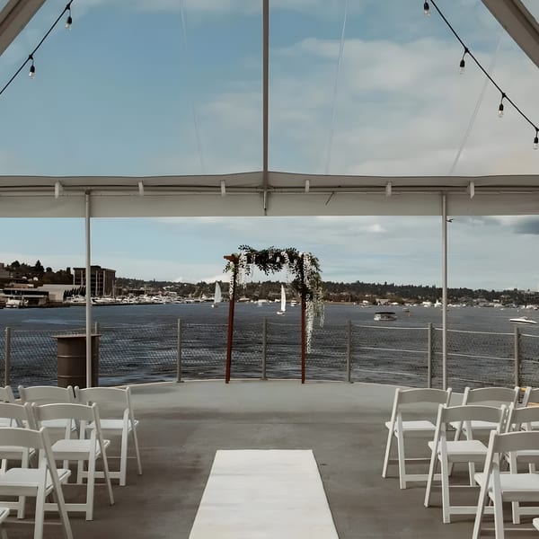 Outdoor wedding ceremony setup with white chairs, floral arch, and waterfront view under a clear tent.