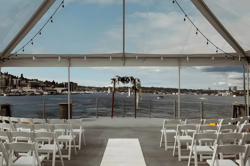 Outdoor wedding ceremony setup with white chairs, floral arch, and waterfront view under a clear tent.