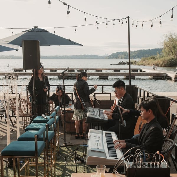 A band performs on a patio overlooking a lake, with string lights overhead and drinks on tables.