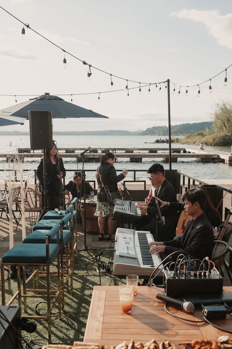 A band performs on a patio overlooking a lake, with string lights overhead and drinks on tables.