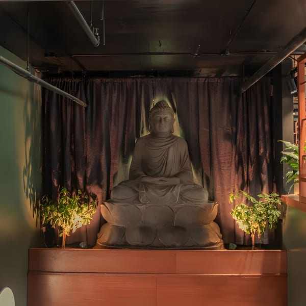 A large Buddha statue sits on a wooden platform, flanked by plants and illuminated by soft lighting.