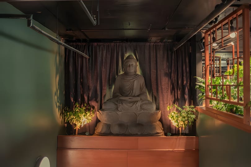A large Buddha statue sits on a wooden platform, flanked by plants and illuminated by soft lighting.