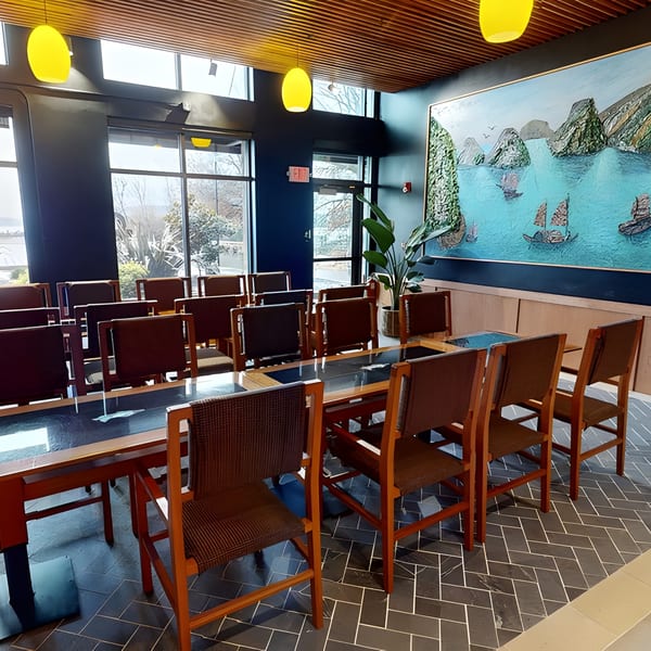 A private dining room with a long table, chairs, large windows, and a vibrant seascape painting.