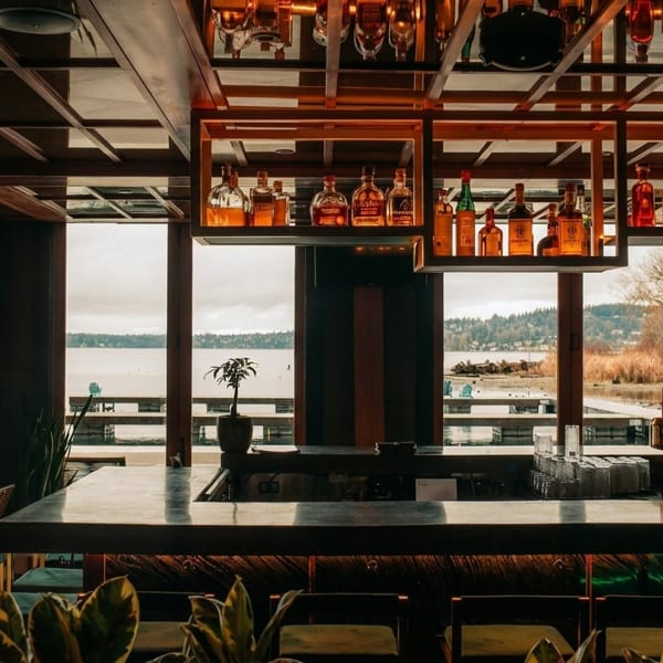 A modern bar with a wooden ceiling, liquor bottles, and large windows overlooking a lake.