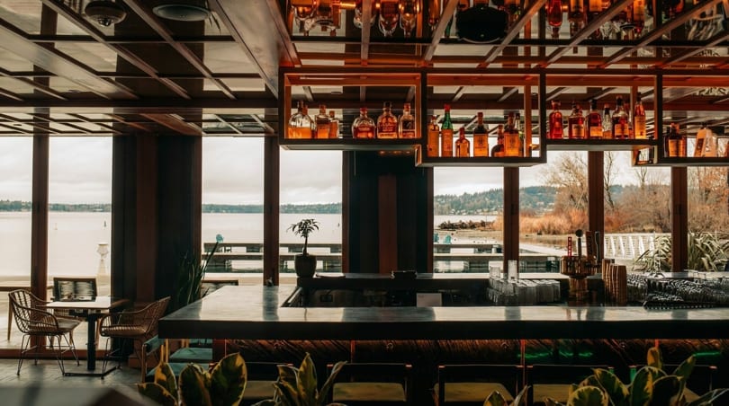 A modern bar with a wooden ceiling, liquor bottles, and large windows overlooking a lake.