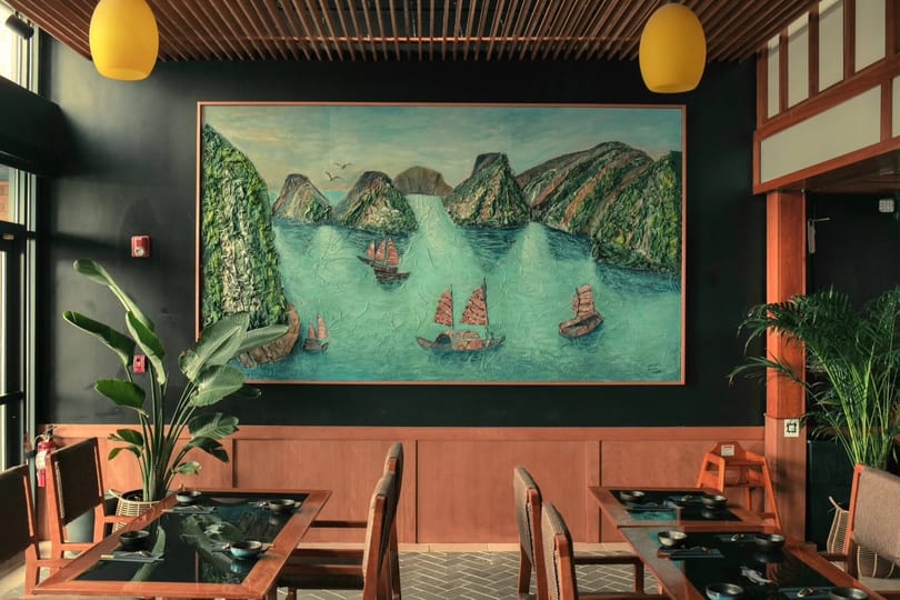Restaurant interior with a large textured painting of boats on water, wooden tables, and plants.