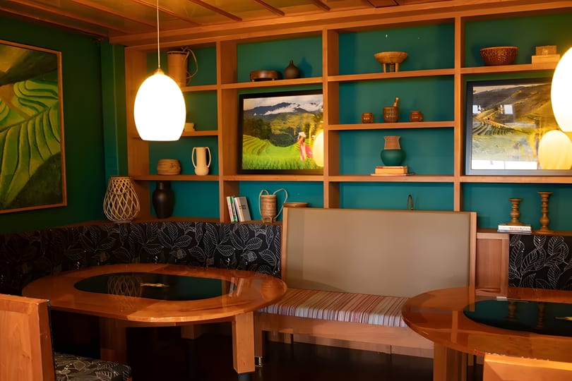 A restaurant interior with green walls, wooden shelves displaying decor, and booth seating with round tables.