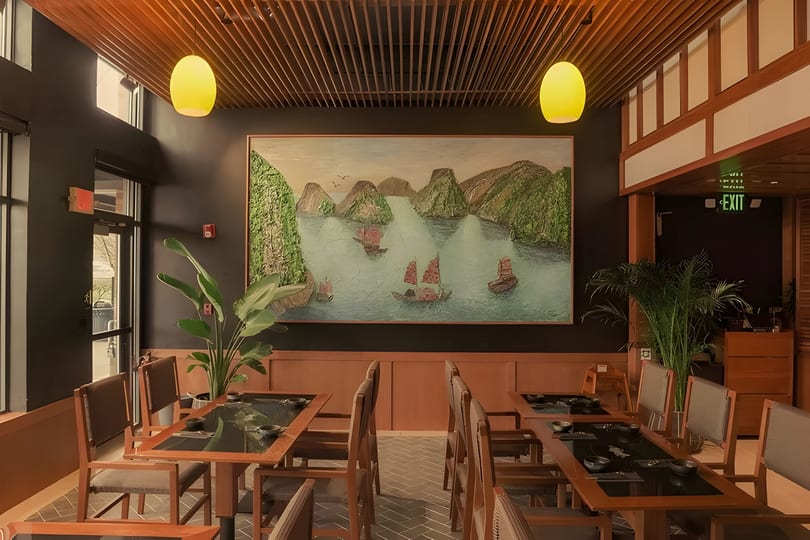 Restaurant interior with wooden tables, chairs, a large mural of boats on water, and yellow pendant lights.