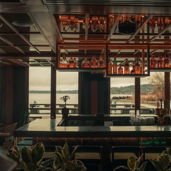 A modern bar with bottles on shelves, overlooking a lake and distant shoreline through large windows.
