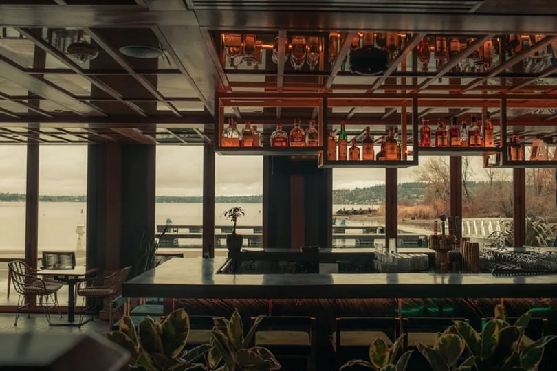 A modern bar with bottles on shelves, overlooking a lake and distant shoreline through large windows.