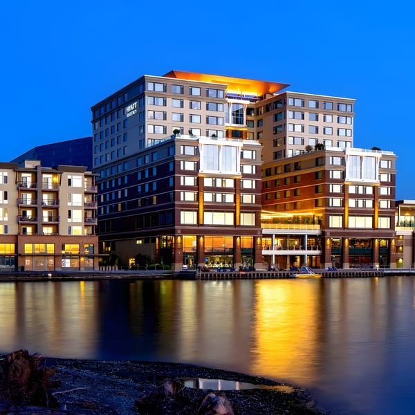 A large, illuminated Hyatt Regency hotel and other buildings line a waterfront at dusk.