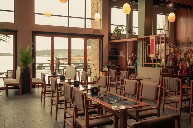 Spacious restaurant dining room with wooden tables and chairs, large windows overlooking a lake.
