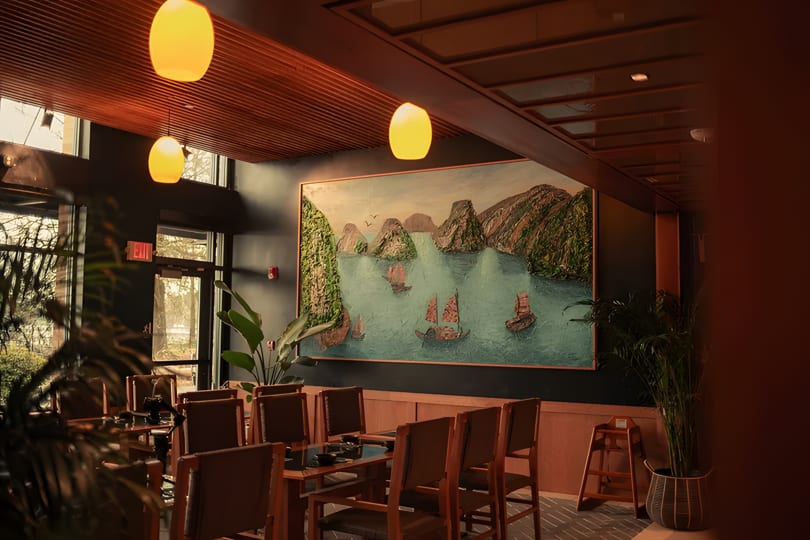 Restaurant interior with a large painting of boats on water, wooden tables, and warm lighting.