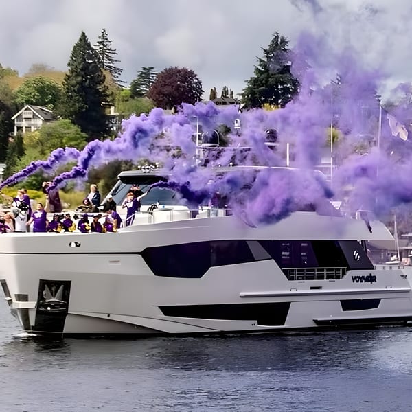 A white yacht with people on board emits purple smoke, cruising on water with houses and trees in the background.