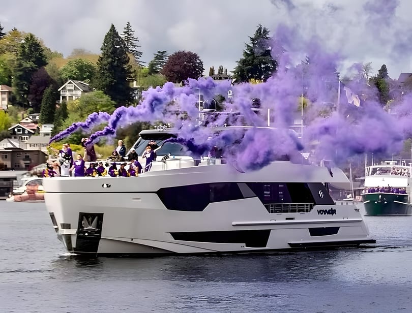 A white yacht with people on board emits purple smoke, cruising on water with houses and trees in the background.