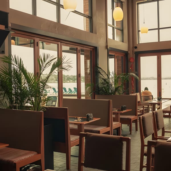 Restaurant interior with wooden tables, brown chairs, potted plants, and large windows overlooking water.