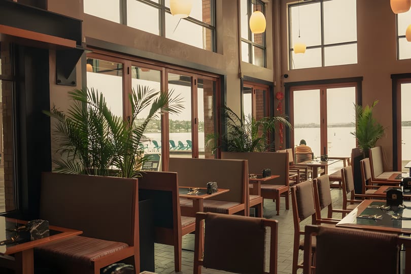Restaurant interior with wooden tables, brown chairs, potted plants, and large windows overlooking water.