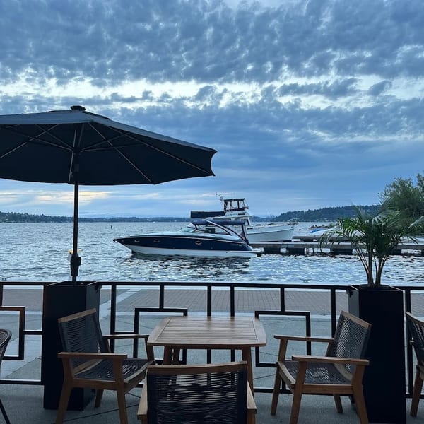 Outdoor dining patio with tables, chairs, and umbrellas overlooking a lake with boats and a cloudy sky.