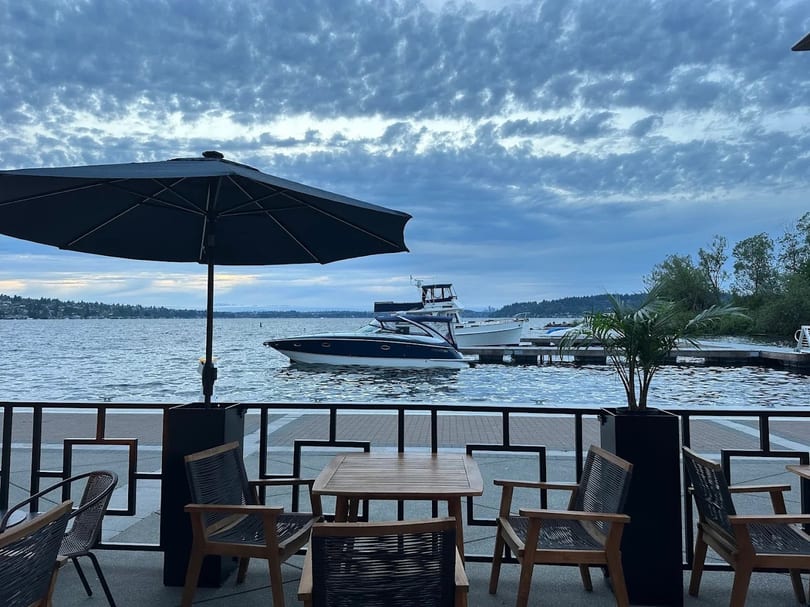 Outdoor dining patio with tables, chairs, and umbrellas overlooking a lake with boats and a cloudy sky.
