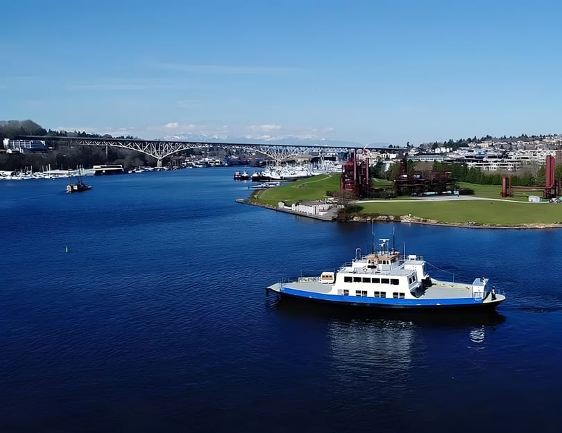 A ferry boat on a river with a bridge, city, and mountains in the background.