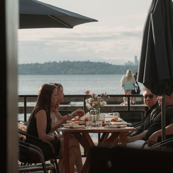 Patrons dine on an outdoor patio overlooking a lake with a distant city skyline.