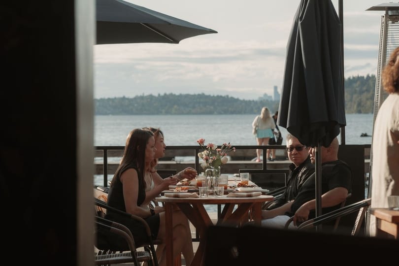 Patrons dine on an outdoor patio overlooking a lake with a distant city skyline.