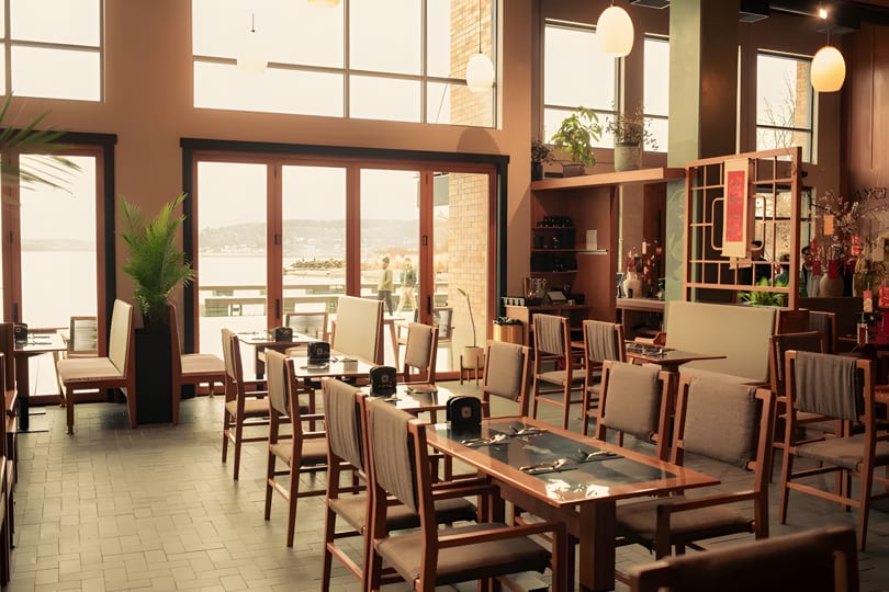A spacious, modern restaurant dining room with wooden tables, chairs, and large windows overlooking a body of water.
