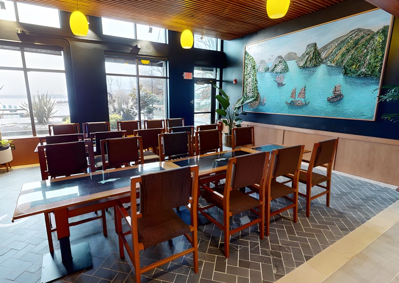 A private dining room with a long table, chairs, large windows, and a vibrant seascape painting.